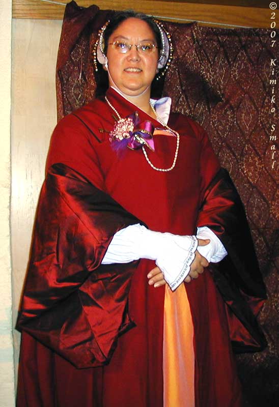 A photo of me, Kimiko, wearing my red wool & silk Tudor gown c.1530s, topped by my white silk French hood.